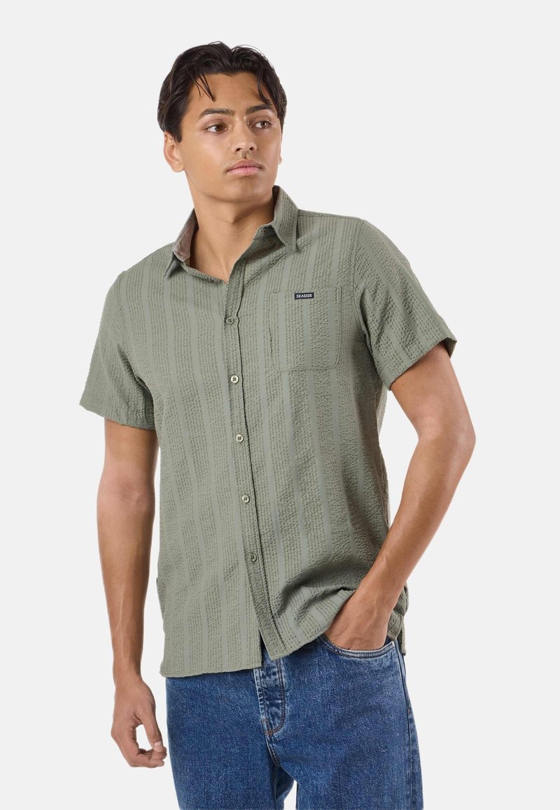 Homme portant une chemise verte à boutons texturée à manches courtes avec une poche poitrine et un jean bleu, debout avec une main dans la poche.