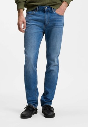 Mann trägt blaue Jeans mit geradem Schnitt, schwarze Schnürschuhe und einen grünen Pullover mit einer Hand in der Tasche.