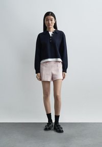 Nainen seisoo yksiväristä seinää vasten, yllään tummansininen napitettu villapaita, valkoinen aluspaidan, vaaleanpunaiset teksturoidut shortsit, mustat sukat ja mustat loaferit.