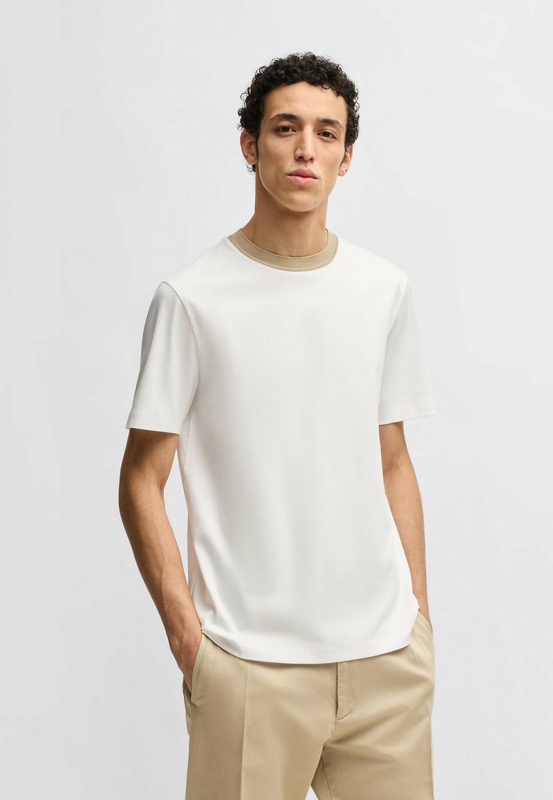 T-shirt bianco a maniche corte con colletto a coste beige, texture liscia e design classico a girocollo; abbinato a pantaloni beige.