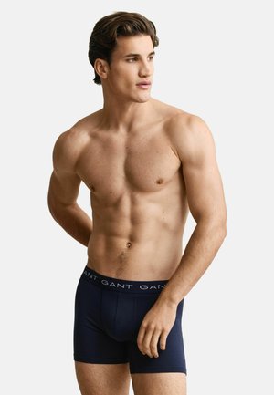 Man zonder shirt met gespierd lichaam, gekleed in marineblauwe boxershorts met "GANT" tailleband, kijkt naar links tegen een effen achtergrond.