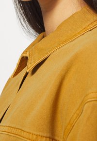 Hořčicově žlutá denimová bunda s špičatým límcem, viditelným šitím a strukturovanou tkaninou. Detailní pohled zaměřený na límec a rameno.