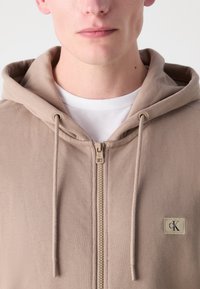 Felpa beige con zip realizzata in tessuto morbido, dotata di cappuccio regolabile e un piccolo patch con il marchio sulla parte sinistra del petto.