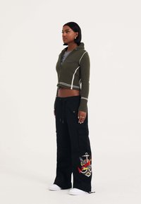 Donkergroen cropped zip-up trui met witte accenten, gecombineerd met zwarte cargobroek met kleurrijke borduursels en witte sneakers.