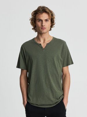 Giovane uomo con capelli biondi ricci che indossa una maglietta Henley verde oliva a maniche corte, in piedi davanti a uno sfondo semplice.