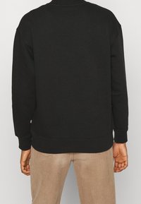 Sudadera negra con cuello alto, puños acanalados y dobladillo. La parte posterior presenta una textura suave y no tiene gráficos ni logos visibles.