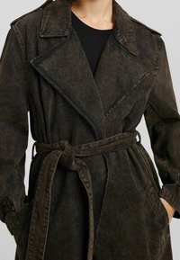 Ciemnobrązowy płaszcz trench wykonany z teksturowanej tkaniny, z paskiem do wiązania, dużymi klapami i dwoma przednimi kieszeniami. Projekt jest luźny i warstwowy.