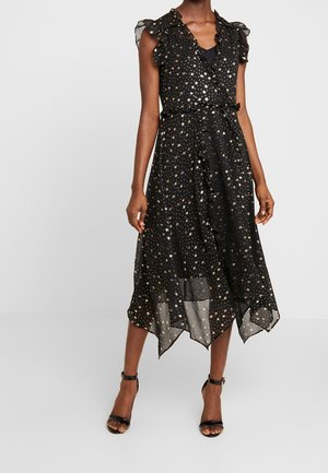 Femme portant une robe midi noire ornée de motifs d'étoiles dorées, avec des manches à volants et un ourlet en mouchoir, associée à des escarpins noirs à bride cheville.