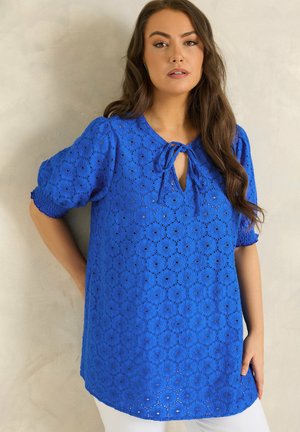 Vrouw draagt een felblauwe oogkantblouse met korte pofmouwen en een keyhole striknek, staand tegen een neutrale muur.