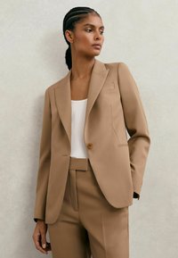 Moteris su pintinėmis plaukais, dėvinti smėlio spalvos pritaikytą kostiumą ir baltą viršutinę dalį, stovinti priešais šviesią tekstūruotą sieną, žiūrinti į šoną.