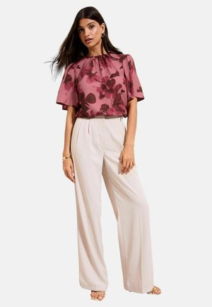 Sieviete valkā brīvi pieguļošu rozā ziedu raksta blūzi ar īsām piedurknēm, kas apvienota ar augstās jostasvietas plašu krēmkrāsas biksēm un atvērtu purngala sandales.