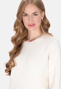 Blonde Frau mit langen, welligen Haaren, die Creolen und einen weißen Strickpullover trägt, blickt mit neutralem Ausdruck direkt in die Kamera.