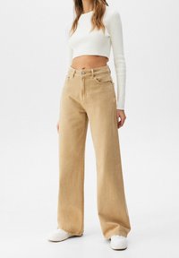 Beige leveled jeans dengan pinggang tinggi dan lima kantong, dipadukan dengan atasan crop putih bertekstur rib dan sepatu sneakers putih.