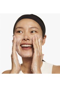 Application d'un nettoyant facial sur le visage souriant d'une personne ; texture gélatineuse avec des bulles abondantes ; peau claire, maquillage minimal et bandeau noir.