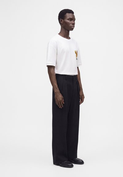 Mann steht in weißem T-Shirt mit kleinem buntem Design auf der Brust, schwarzen hoch taillierten, weiten Hosen und schwarzen Schuhen vor weißem Hintergrund.