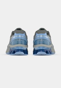 Ténis desportivos com parte superior cinzenta com detalhes azuis, malha texturada e uma sola espessa e distintiva com gel para amortecimento.