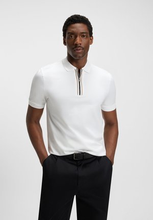 White polo shirt with short sleeves, zippered placket featuring a black and brown accent. Worn with black trousers, smooth fabric texture.