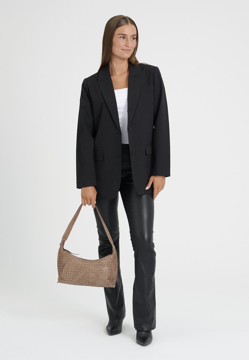 Femme portant un blazer noir et un pantalon en cuir, tenant un sac à main tressé marron sur un fond blanc uni.
