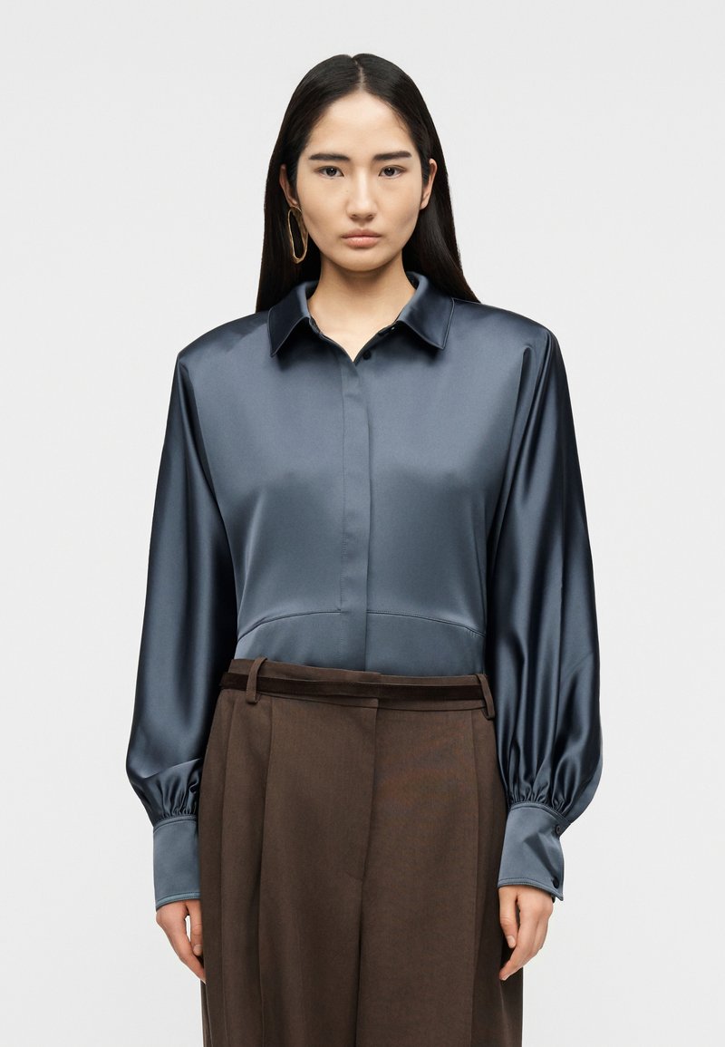 Γυναίκα με μακριά σκουρόχρωμα μαλλιά φοράει μπλε σατέν πουκάμισο με φουσκωτά μανίκια και καφέ παντελόνι με ψηλή μέση, στέκεται μπροστά σε απλό φόντο.