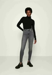 Mulher com cabelo curto e encaracolado, vestindo um top de gola alta preto, calças skinny cinzas de cintura alta e sapatos de plataforma pretos, de pé contra um fundo simples.