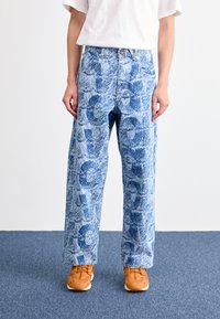 Personne portant un jean bleu clair avec des motifs abstraits bleu foncé, un t-shirt blanc et des baskets orange, se tenant sur un tapis bleu.