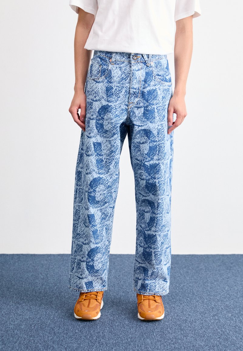 Personne portant un jean bleu clair avec des motifs abstraits bleu foncé, un t-shirt blanc et des baskets orange, se tenant sur un tapis bleu.