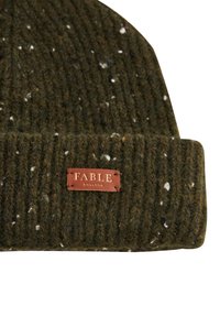 Zaļš adīts cepure ar ribotu tekstūru, kas bagātināta ar baltu un zemes toņu plankumiem, un taisnstūrveida brūna ādas etiķete ar uzrakstu "FABLE ENGLAND".