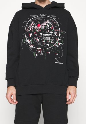 Sweat à capuche noir avec un motif de montagne abstraite, un soleil rouge et la mention « A crisp layer of madness », porté par une personne du torse jusqu'à mi-cuisse.