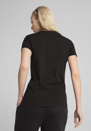 Blonde Frau mit goldenen Creolen, schwarzem kurzärmligem T-Shirt und schwarzer Hose, steht mit dem Rücken zur Kamera vor einem schlichten grauen Hintergrund.