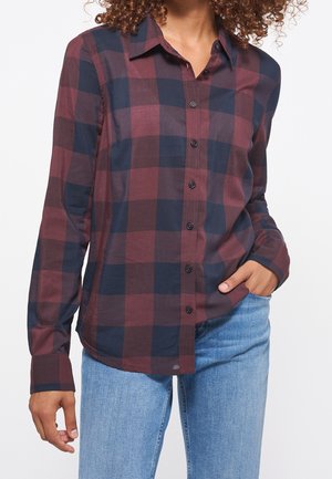 Kvinde iført en ternet skjorte i vinrød og marineblå med knapper og lange ærmer samt lyseblå jeans, med den ene hånd i lommen.