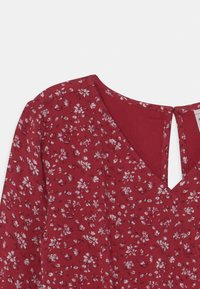 Blusa roja con estampado floral, con escote en V y una apertura en forma de llavero en la parte posterior. Presenta una tela texturizada con pequeños patrones de flores blancas y rosas.