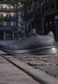 Black athletic sneaker with textured sole and three side stripes, placed on cobblestone street in front of blurred urban buildings.