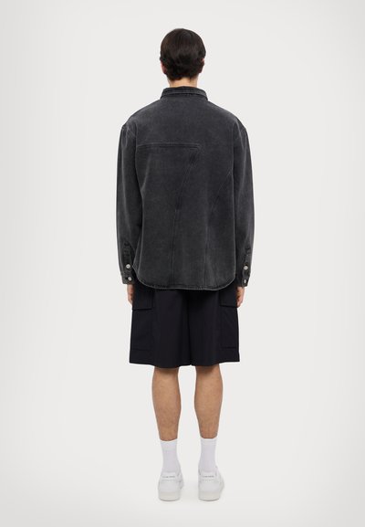 Zwart denim shirtjack met een losse pasvorm, lange mouwen en knoopaccenten, gecombineerd met zwarte cargoshorts en witte sneakers.