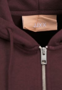 Felpa zip-up color bordeaux con zip argentata e un'etichetta beige con il testo "JJXX." Tessuto morbido e strutturato, cappuccio con lacci.