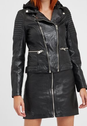 Mujer con una chaqueta de cuero negro con mangas acolchadas y cremalleras plateadas, combinada con una falda de cuero negro que tiene una cremallera frontal.