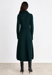 Knielanges, dunkelgrünes Strickkleid mit hohem Kragen, taillierter Taille, langen Ärmeln und subtiler Rückennaht. Getragen mit schwarzen, kniehohen Stiefeln.