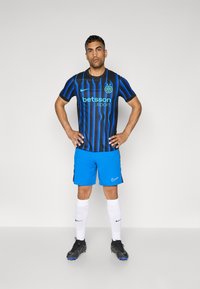 Maillot de football rayé noir et bleu avec un logo, associé à un short bleu et des chaussettes blanches. Baskets noires avec des accents bleus.