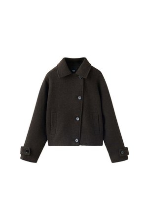 Jeune femme debout portant une veste foncée boutonnée, un top court blanc, un jean large bleu clair, des chaussures marron, tenant un vêtement rouge dans une main.