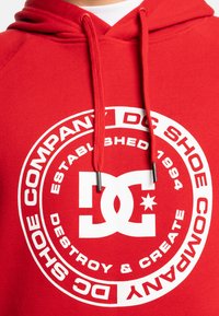 Red hoodie with a large white circular logo. Text includes "DC SHOES COMPANY" and "ESTABLISHED 1994." Features drawstrings with metal tips.