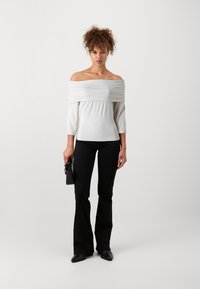 Hvit off-shoulder topp med trekvartlange ermer, sammen med svarte vide bukser og svart håndveske, stående mot en lys bakgrunn.