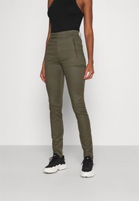 Olivgrön figurnära byxor med en mjuk textur, utrustade med en dragkedjeficka och hög midja, tillsammans med svarta sneakers och en svart topp.