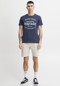 Marineblå bomulls t-skjorte med "Blend Denim Original Vintage" grafikk i lysegrønn og hvit. Kombinert med beige shorts og svarte joggesko.