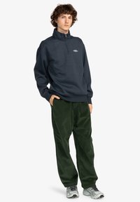 Navy half-zip pullover with a relaxed fit and ribbed hem, paired with dark green corduroy pants. Features a simple logo on the chest.