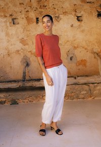 Femme souriante, portant un haut en maille à manches courtes de couleur rouille, un pantalon blanc taille haute, des talons ouverts noirs, debout près d'un mur beige texturé.