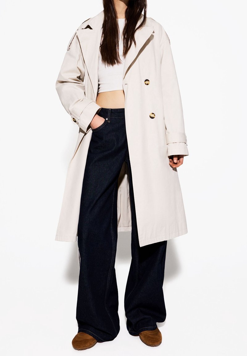 Femme porte un trench beige sur un crop top blanc, un jean large foncé et des chaussures marron, debout avec une main dans la poche sur fond blanc.