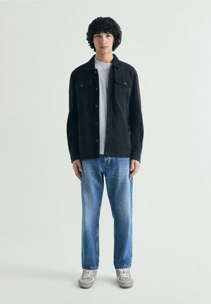 Jeune homme aux cheveux bouclés portant une veste noire boutonnée, un T-shirt blanc, un jean bleu et des baskets grises, debout devant un fond uni.