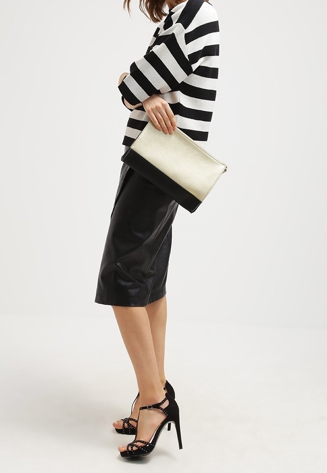 Femme en pull rayé noir et blanc, jupe en cuir noir, sandales à talons hauts noires, tenant une pochette noir et blanc.