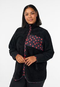 Schwarze Fleecejacke mit hohem Kragen und Paneel mit rotem Kirschmuster. Verfügt über einen Reißverschluss und kontrastierende rote Besätze an Taschen und Bündchen.