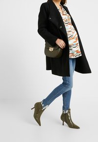 Svart dubbelknäppt kappa över en mönstrad topp, blå tighta jeans och olivgröna spetsiga ankle boots med strukturerad yta.