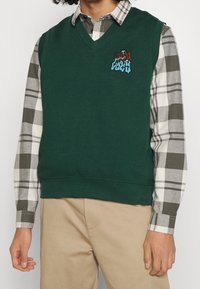 Mörkgrön ärmlös tröja med V-ringning, prydd med en texturerad broderad design på bröstet, lagd över en rutig skjorta.